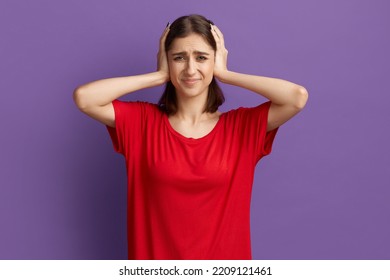 Turn The Volume Down. Dissatisfied Frustrated Young Pretty Brunette Girl In Red T-shirt Covers Ears With Palms. Being In Rage Looks Dissatisfied And Unhappy. Posing Over Purple Wall