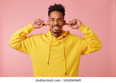 Turn Volume, Please! Annoyed Young African American Man Pluggs Ears And Clenches Teeth, Dissatisfied With Loud Music, Wears In Yellow Hoodie, Stands Over Pink Background With Closed Eyes.