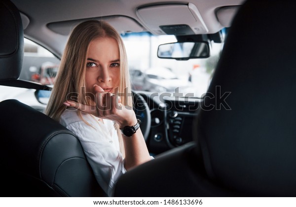 Turned back to\
the cameramen at backseat. Beautiful blonde girl sitting in the new\
car with modern black\
interior.
