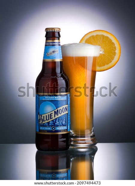 TURNERS FALLS, UNITED STATES - Dec 01, 2021: A vertical\
closeup of the Blue Moon beer bottle and a full glass of beer on a\
reflective surface 