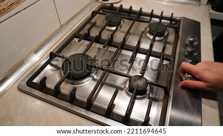 Turns off or turns on the gas on the stove. Blue flame of fire on black burners. The man's hand turns off or turns on the gas. Black grilles on a steel plate. White tiles on the walls. Cold winter