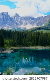 Turquoise alpine lake reflecting a dramatic rocky mountain range and dense evergreen forest beneath a partly cloudy sky.