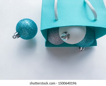 Turquoise (blue) paper gift bag, inside of which are visible shiny Christmas balls of blue, silver and white colors. One turquoise ball lies on the left. White background, copy space