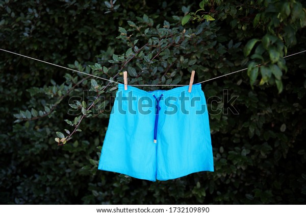 Men’s turquoise blue swimming costume\
hanging out to dry on string clothing line, secured with two wooden\
pegs, hanging against dark leafy\
background