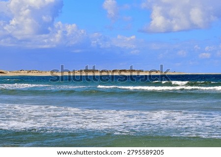 Turquoise and deep blue waves roll toward a pale sandy shoreline, their white foam catching the light as low dunes with sparse vegetation rise behind them. The scene blends vivid ocean colour with the soft, natural textures of a calm coastal landscape.