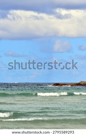 Turquoise and deep blue waves roll toward the shore, their white foam catching the light as distant rocky formations rise from the sea beneath a bright sky scattered with soft clouds. The scene blends gentle ocean motion with a calm, open coastal horizon.