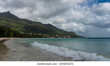 Turquoise ocean waves roll onto the sandy beach and foam.  A green hill against the sky and cumulus clouds. Seychelles. Mahe. Beau Vallon - Shutterstock ID 2193355471