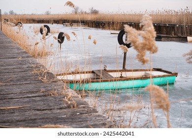 A turquoise wooden rowboat moored in winter on the frozen lake amidst the reeds. A wooden dock surrounds the harbor. - Powered by Shutterstock