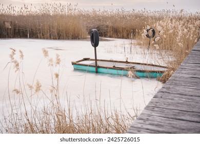 A turquoise wooden rowboat moored in winter on the frozen lake amidst the reeds. Wooden pier. - Powered by Shutterstock