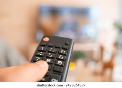 TV Entertainment Concept. A hand firmly grasps the TV remote control, directing it towards the television screen, while the background, featuring the TV and living room remains blurred - Powered by Shutterstock