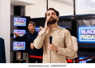 TV reporter live broadcasting talking about Black Friday lines in local shopping mall, holding microphone and looking at camera. Young man correspondent covering news about sales season in retail