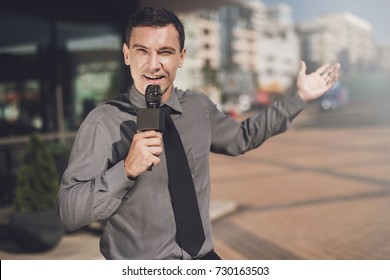 TV Reporter At Work. The Journalist Is Smiling And Is Reporting On The Street In Front Of The Cafe And Apartment Buildings