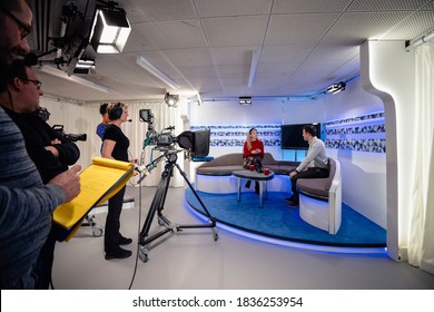 A TV Show Being Filmed In A Studio. The Presenters Are Sitting On The Studio Sofa And Talking To Each Other While The Camera Crew Films Them With Film Cameras.