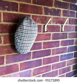 A Tweed Flat Cap Hanging On A Coat Hook Against A Brick Wall In A Hallway.