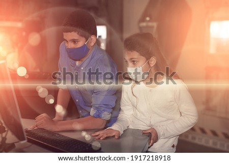 Tween girl and boy in protective masks using computer while trying to find solution of conundrum in quest room. New norms in pandemic, precaution for viral infections. Toned image