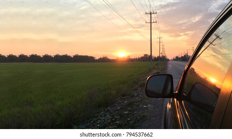 Twilight Scene Of The Journey.The Reflection Of Sunset Was Shown On Black SUV Mirror,green Rice Field And Golden Sky.The Charming Countryside Climate And The Long Way Ahead. In The Middle Way Concept