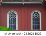 Twin Arched Windows in a church with Intricate White Panes and Blue Glass Details, Set into a Reddish-Brown Building Facade Beneath a Traditional Tiled Roof and Gutter System