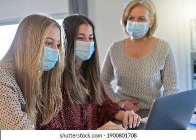 Twin Sisters And Their Teacher Wearing Protective Mask And Working Their Lesson  On  A Laptop At Home During Lockdown Due To Covid-19