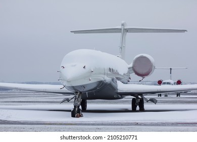 A Twin-engine Jet Plane Covered With Snow Stands At The Airport In The Parking Lot