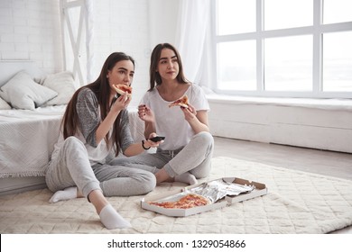 Twins have nice weekend. Sisters eating pizza when watching TV while sits on the floor of beautiful bedroom at daytime.