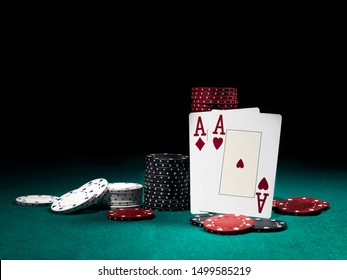Two aces hearts and diamonds standing leaning on chips piles, some of them laying nearby on green cover of playing table. Black background. Close-up.