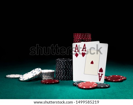 Two aces hearts and diamonds standing leaning on chips piles, some of them laying nearby on green cover of playing table. Black background. Close-up.