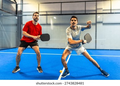 Two active men engaging in a pickleball match on a blue indoor court, concentrating on hitting the ball with their paddles during an athletic competition