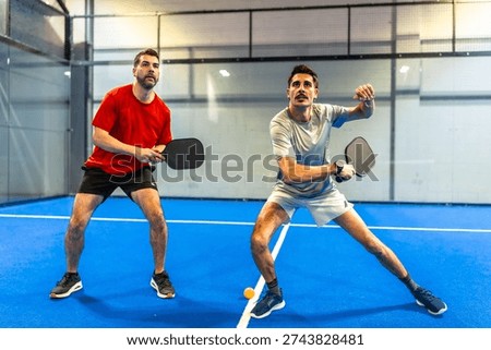 Two active men engaging in a pickleball match on a blue indoor court, concentrating on hitting the ball with their paddles during an athletic competition