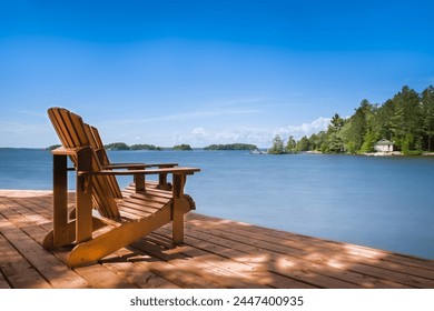 Two Adirondack chairs grace a wooden dock on a sunny summer morning in Muskoka, Ontario, Canada, overlooking the tranquil lake. A lone cottage peeks through the trees on the opposite shore. - Powered by Shutterstock