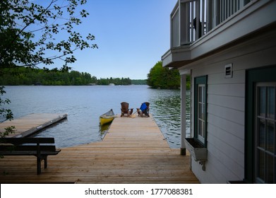Two Adirondack chairs on a cottage wooden dock facing the blue water of a lake in Muskoka, Ontario Canada. A yellow canoe is tied to the dock. Life jacket and oars are visible near the chairs. 