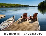 Two Adirondack chairs on a wooden dock facing the blue water of a lake in Muskoka, Ontario Canada. Life jackets are visible near the chairs. A canoe is tied to the pier, paddles are stored inside.