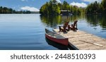 Two Adirondack chairs on a wooden dock overlook the tranquil blue waters of a Muskoka lake in Ontario, Canada. Nearby, a red canoe is tied to the pier, with life jackets and a paddle.