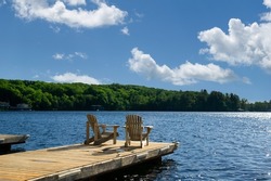 Two Adirondack Chairs On A Wooden Dock Facing A Lake In Muskoka, Ontario Canada During A Sunny Summer Morning. Cottages Are Nested Between Trees Across The Water.
