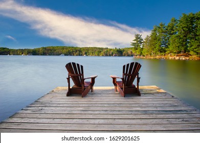 Two Adirondack chairs on a wooden dock facing a calm blue lake. Cottages nestled between green trees are visible across the water.