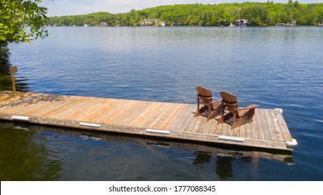 Two Adirondack chairs on a wooden dock overlooking a calm lake. Cottages nestled between green trees are visible across the water. Drone capture.