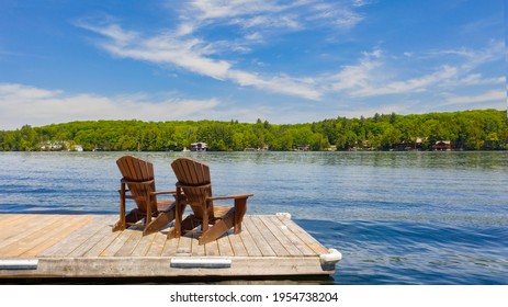 Two Adirondack chairs on a wooden dock facing a lake in Muskoka, Ontario Canada during a sunny summer morning. Cottages are nested between trees across the water.