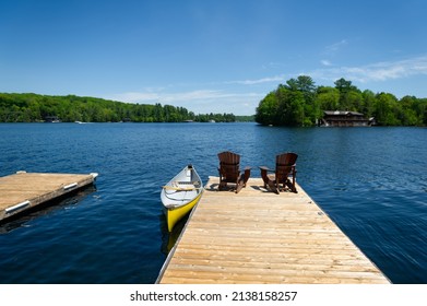 Two Adirondack chairs on a wooden dock facing the blue water of a lake in Muskoka, Ontario Canada. A yellow canoe is tied to the pier. - Powered by Shutterstock