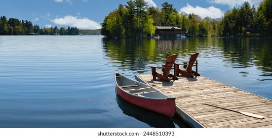 Two Adirondack chairs on a wooden dock overlook the tranquil blue waters of a Muskoka lake in Ontario, Canada. Nearby, a red canoe is tied to the pier, with life jackets and a paddle.