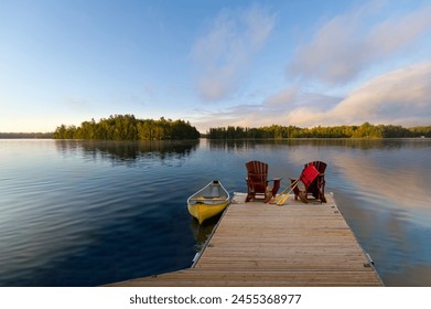 Two Adirondack chairs on a wooden dock overlook the calm lake. A life jacket and paddles hang from one chair, while a vibrant yellow canoe is tied nearby. Across the water, cottages are visible. - Powered by Shutterstock