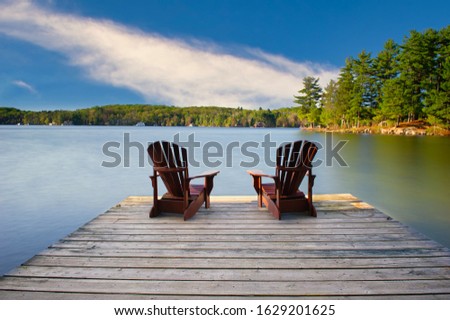 Two Adirondack chairs on a wooden dock facing a calm blue lake. Cottages nestled between green trees are visible across the water.