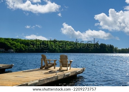 Two Adirondack chairs on a wooden dock facing a lake in Muskoka, Ontario Canada during a sunny summer morning. Cottages are nested between trees across the water.