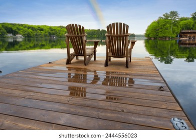 Two Adirondack chairs are positioned on a wet wooden pier, offering a tranquil view of a Muskoka lake. It's early morning at the cottage, with a vibrant rainbow adorning the sky. - Powered by Shutterstock