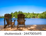 Two Adirondack chairs sit on a wooden dock, overlooking a tranquil lake. On the opposite shore, a white cottage is nestled among lush green trees, with a boat docked in front.