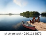 Two Adirondack chairs sit on a wooden dock, gazing at the calm Muskoka lake. A red canoe, life jackets, and a paddle rest nearby, while fluffy clouds and lush green trees reflect on the water.