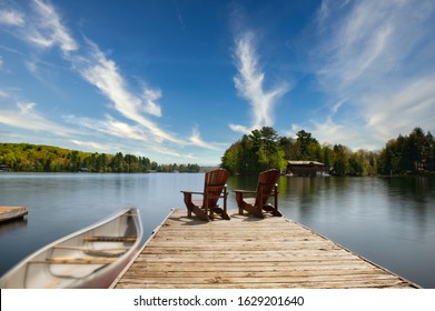 Two Adirondack chairs sit on a wooden dock facing the blue waters of a calm lake. A canoe is tied to the dock. In the background there's a brown cottage nestled between trees. 