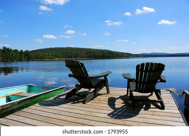 Two Adirondack Wooden Chairs On Dock Facing A Blue Lake With Clouds Reflections
