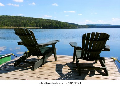 Two Adirondack Wooden Chairs On Dock Facing A Blue Lake With Clouds Reflections