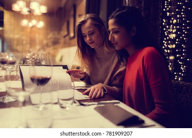 Two Adolescent Women Sitting In A Fancy Restourant, Going Through The Menue, While Drinking Some Red Wine, Talking, Gossiping And Unwrapping Their Christmass Presents And Looking At Their Phones. 