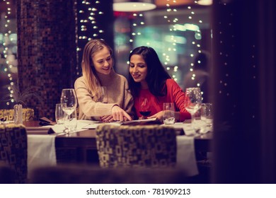 Two Adolescent Women Sitting In A Fancy Restourant, Going Through The Menue, While Drinking Some Red Wine, Talking, Gossiping And Unwrapping Their Christmass Presents And Looking At Their Phones. 