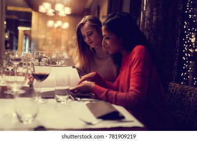 Two Adolescent Women Sitting In A Fancy Restourant, Going Through The Menue, While Drinking Some Red Wine, Talking, Gossiping And Unwrapping Their Christmass Presents And Looking At Their Phones. 